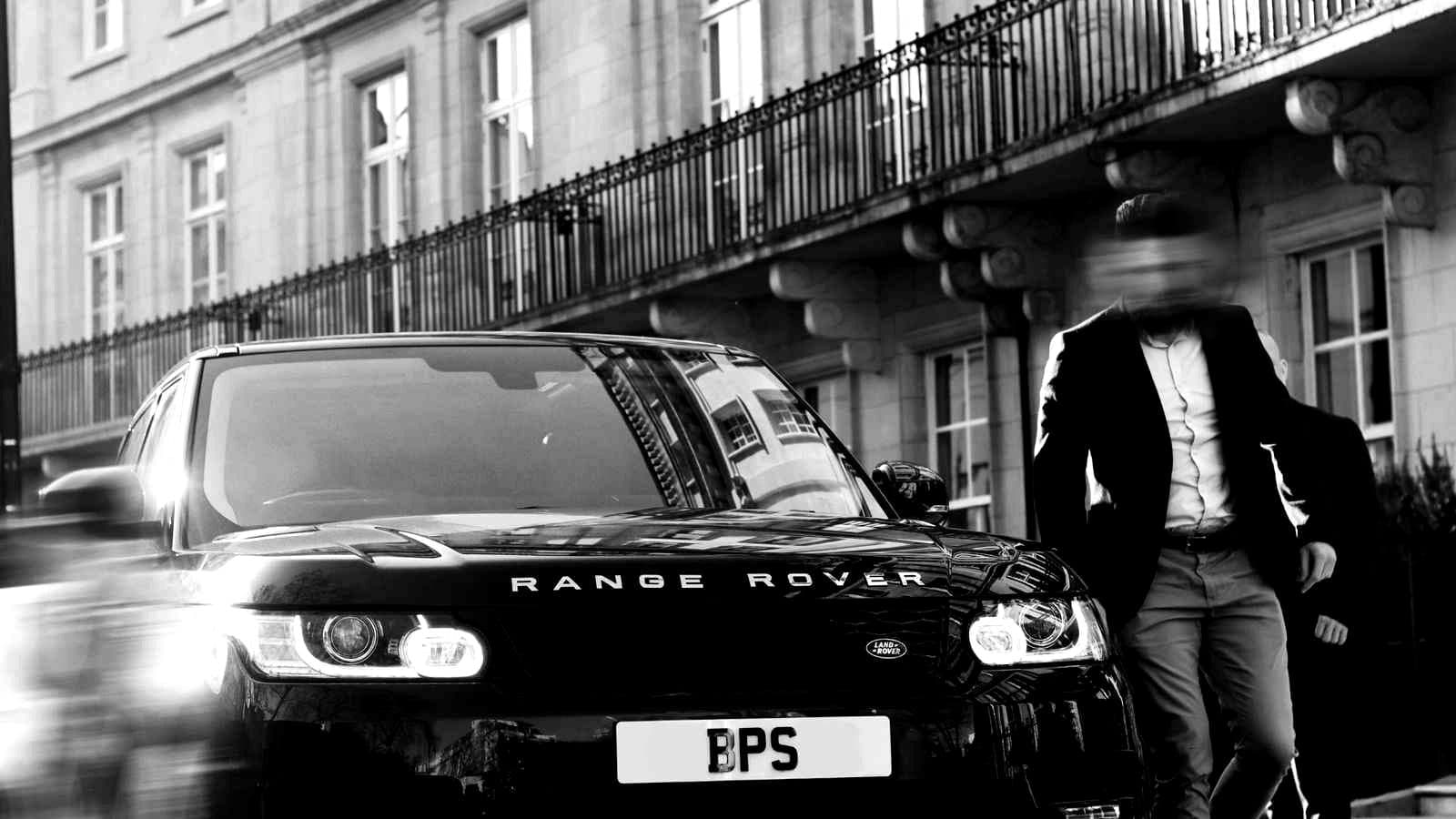 A black BPS Range Rover waiting outside a white stucco terrace, an operative stepping out beside it