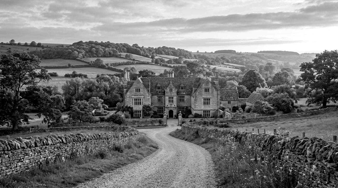 A honey coloured stone manor house at the end of a gravel lane lined with dry stone walls, rolling fields beyond