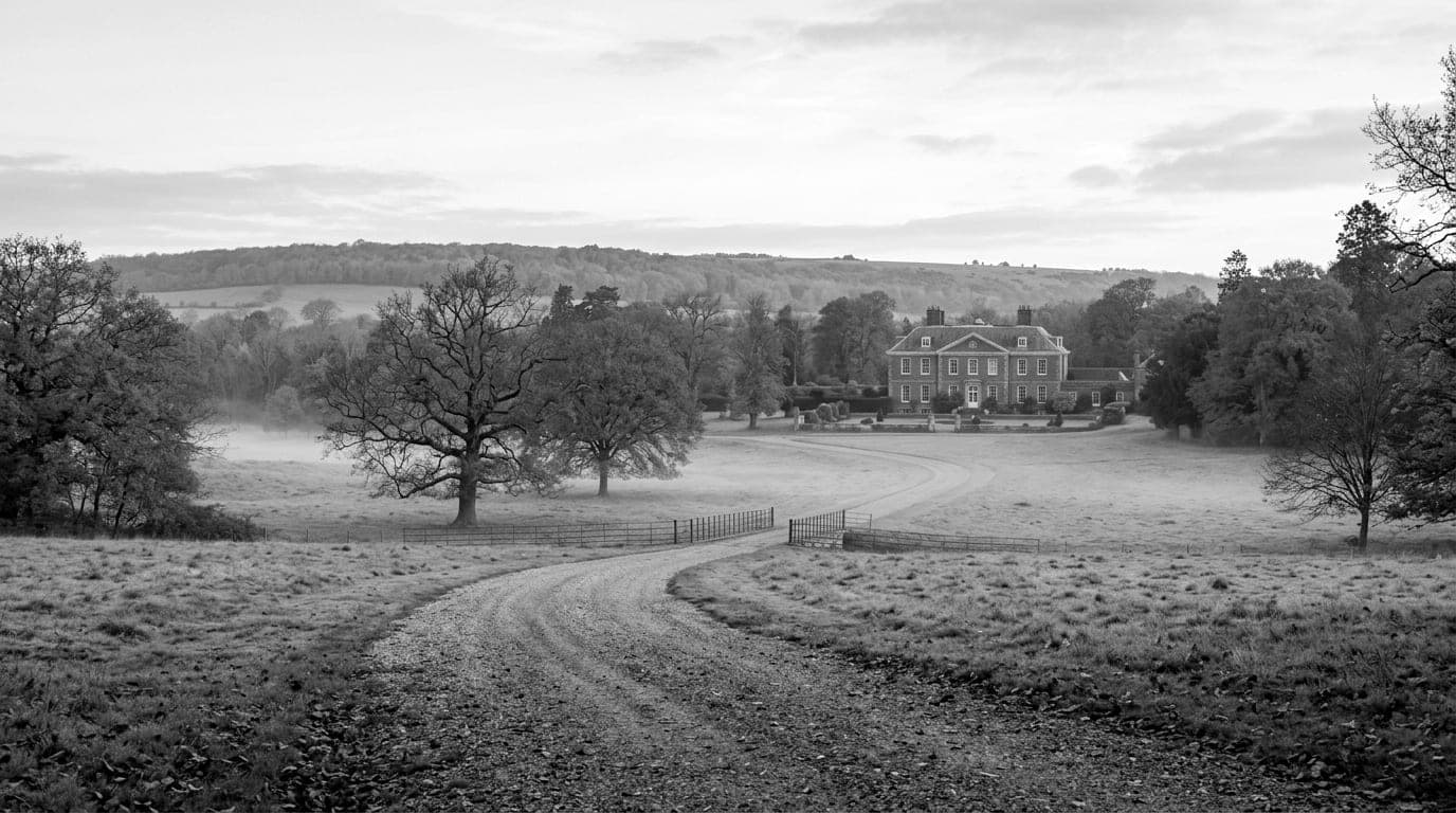 A gravel drive curving through misty parkland towards a red brick country house, a wooded ridge behind