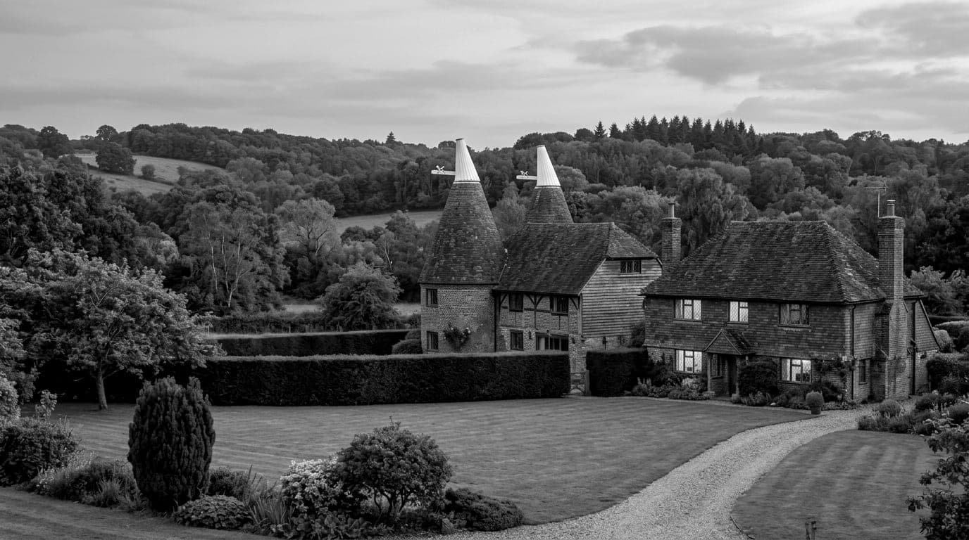 Two converted oast houses beside a tile hung farmhouse, a lawn and clipped hedge in front, wooded hills behind