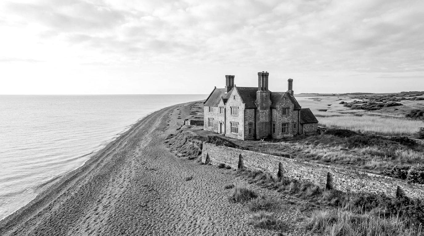 A flint and brick house with tall chimneys behind a low wall above a long shingle beach, reed marsh and heath beyond