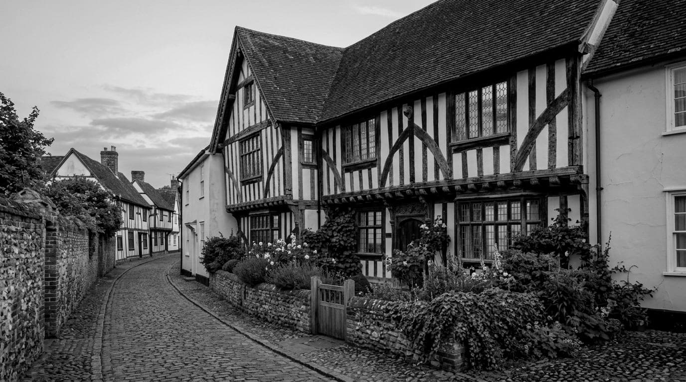 A timber-framed house with leaded windows lit at dusk behind a low flint wall and garden, on a cobbled lane of old houses