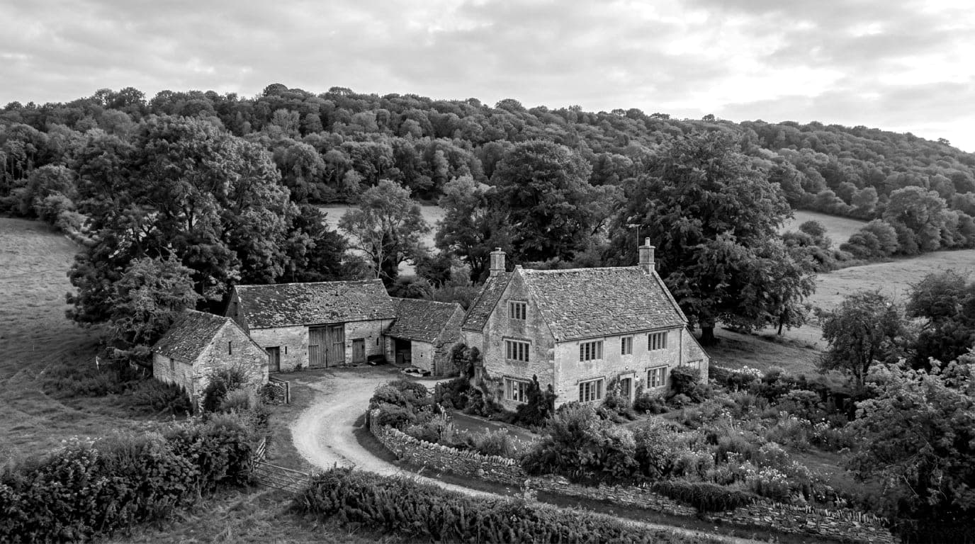A pale stone farmhouse with a stone tiled roof beside old stone barns and a walled garden, a wooded escarpment behind