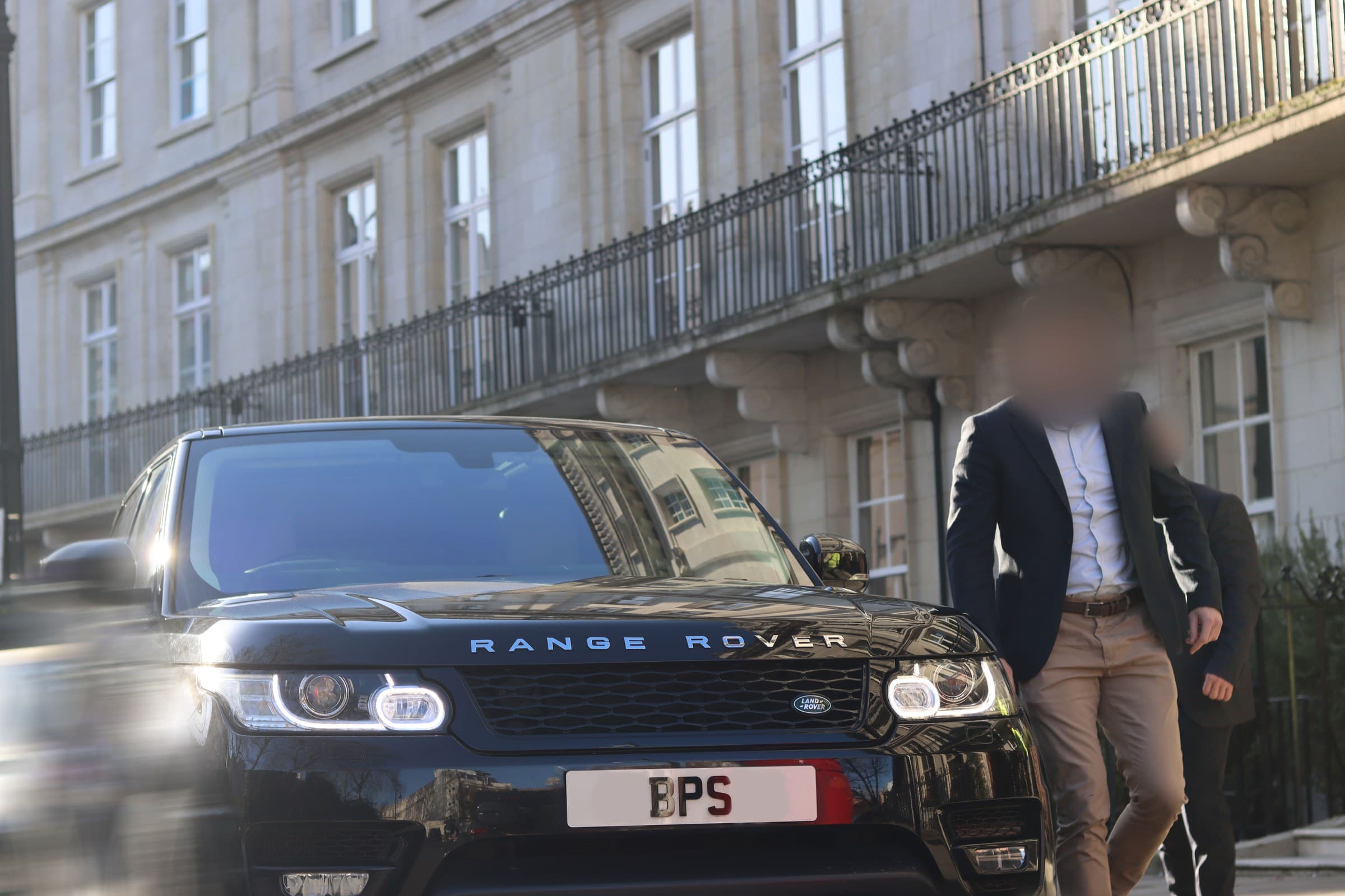 Two men in jackets walking beside the front of a black BPS Range Rover on a street of pale stone terraces with iron balconies