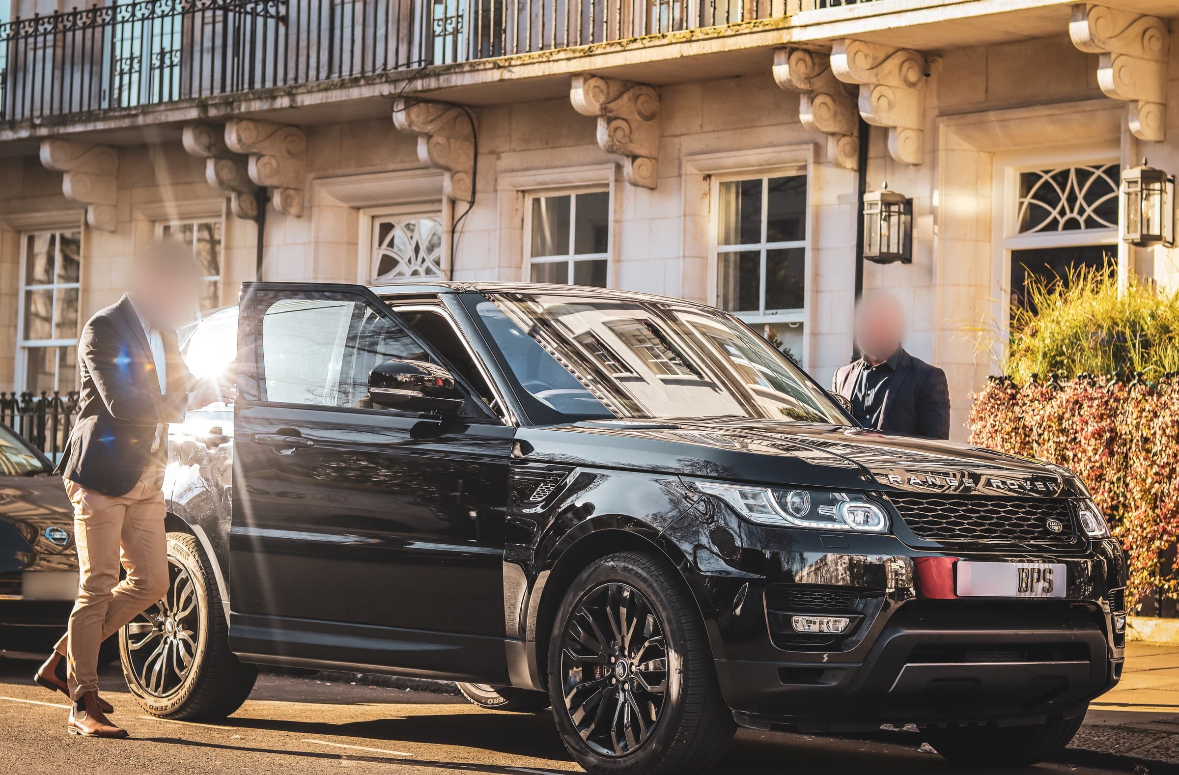 A black BPS Range Rover at the kerb outside a stone building with carved brackets and iron balconies, one man at its open door and another standing at the front of the car