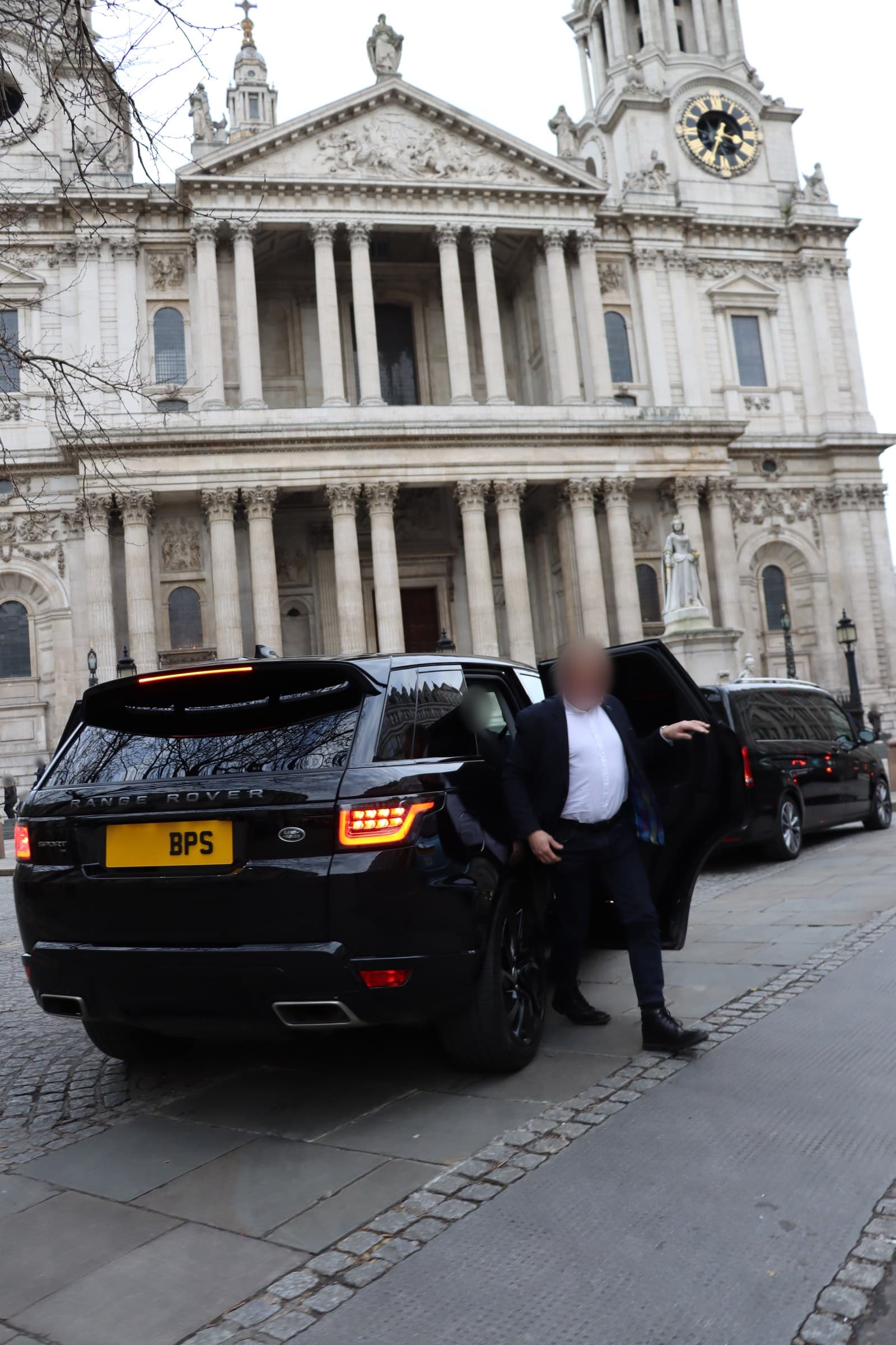 A man in a dark suit stepping out of the rear door of a black BPS Range Rover in front of a columned stone portico, a second black vehicle parked beyond it