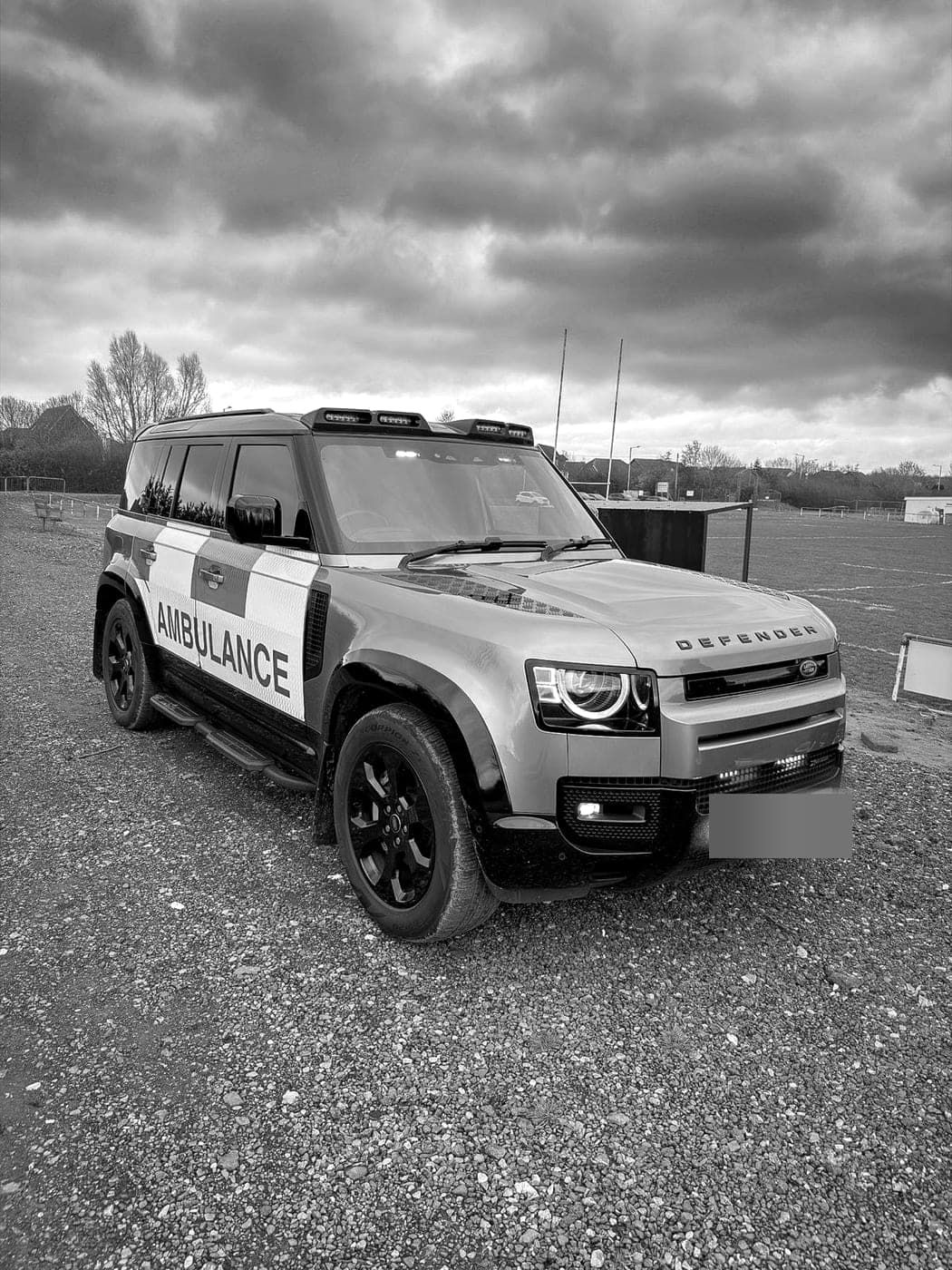 A BPS Land Rover Defender in ambulance markings parked beside a playing field, its number plate blurred