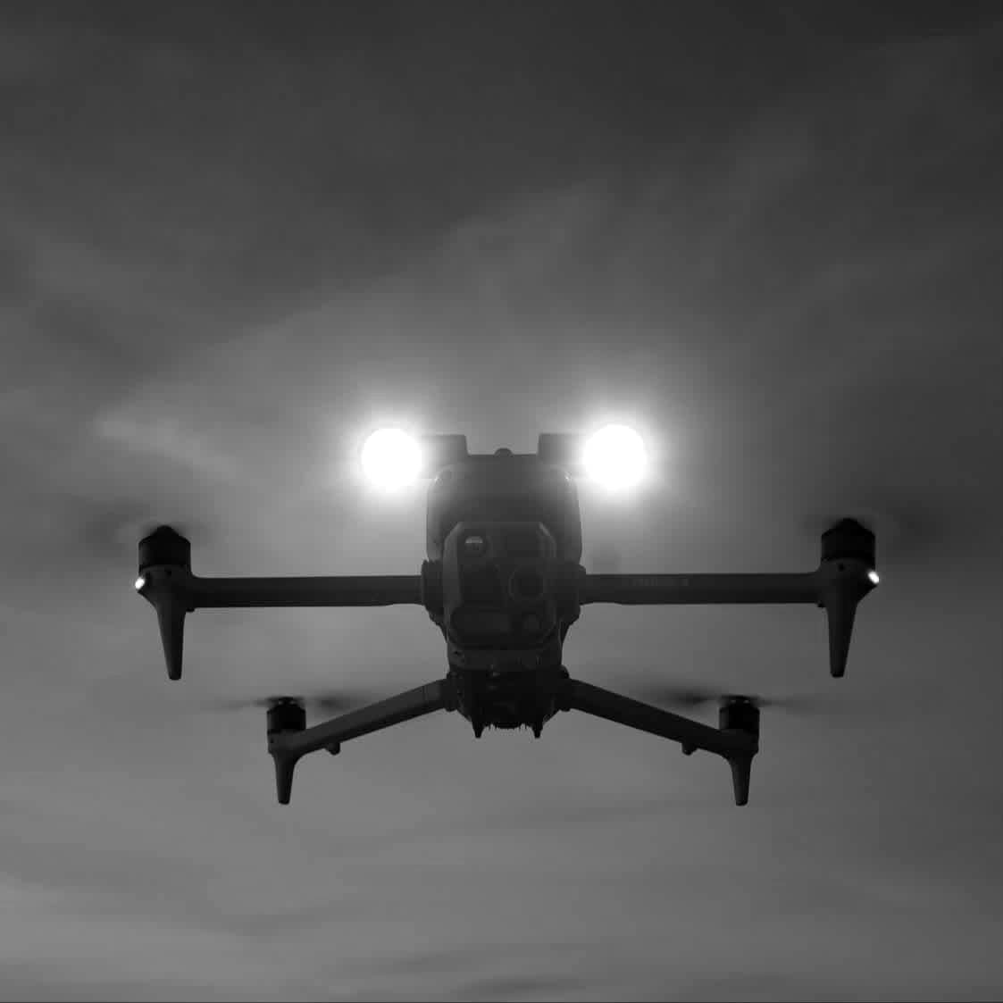 A DJI Matrice drone in flight at dusk, its two spotlights on