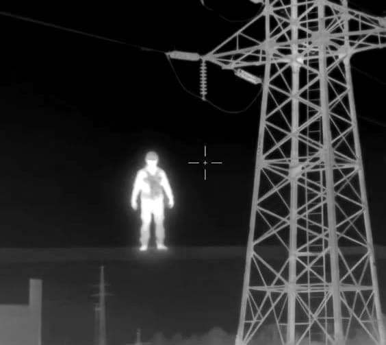 A thermal camera view at night: a person shows up bright against the dark beside an electricity pylon, a crosshair on the image