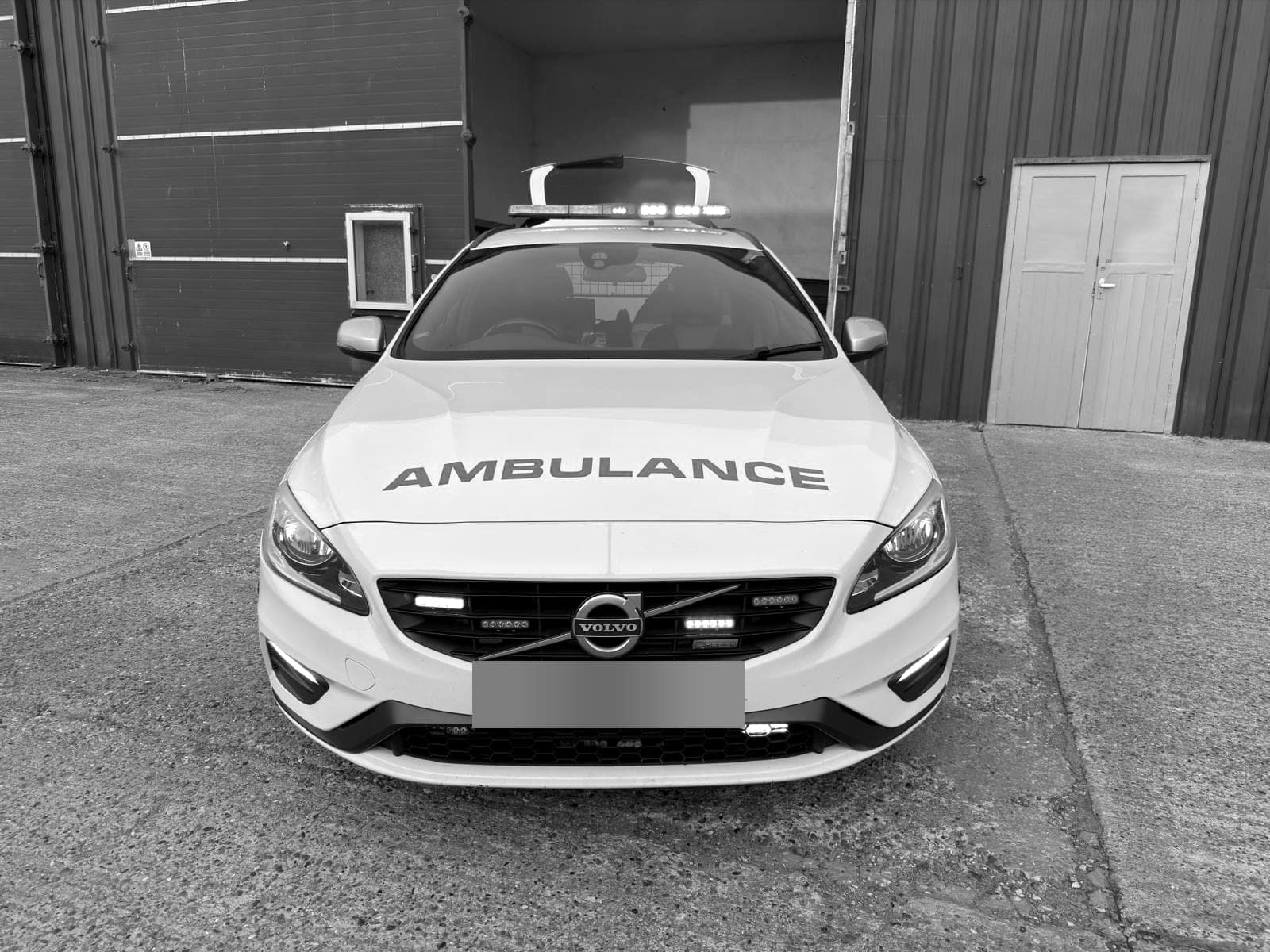 A BPS ambulance car, a Volvo estate in ambulance markings with its lights on, outside a barn, its number plate blurred