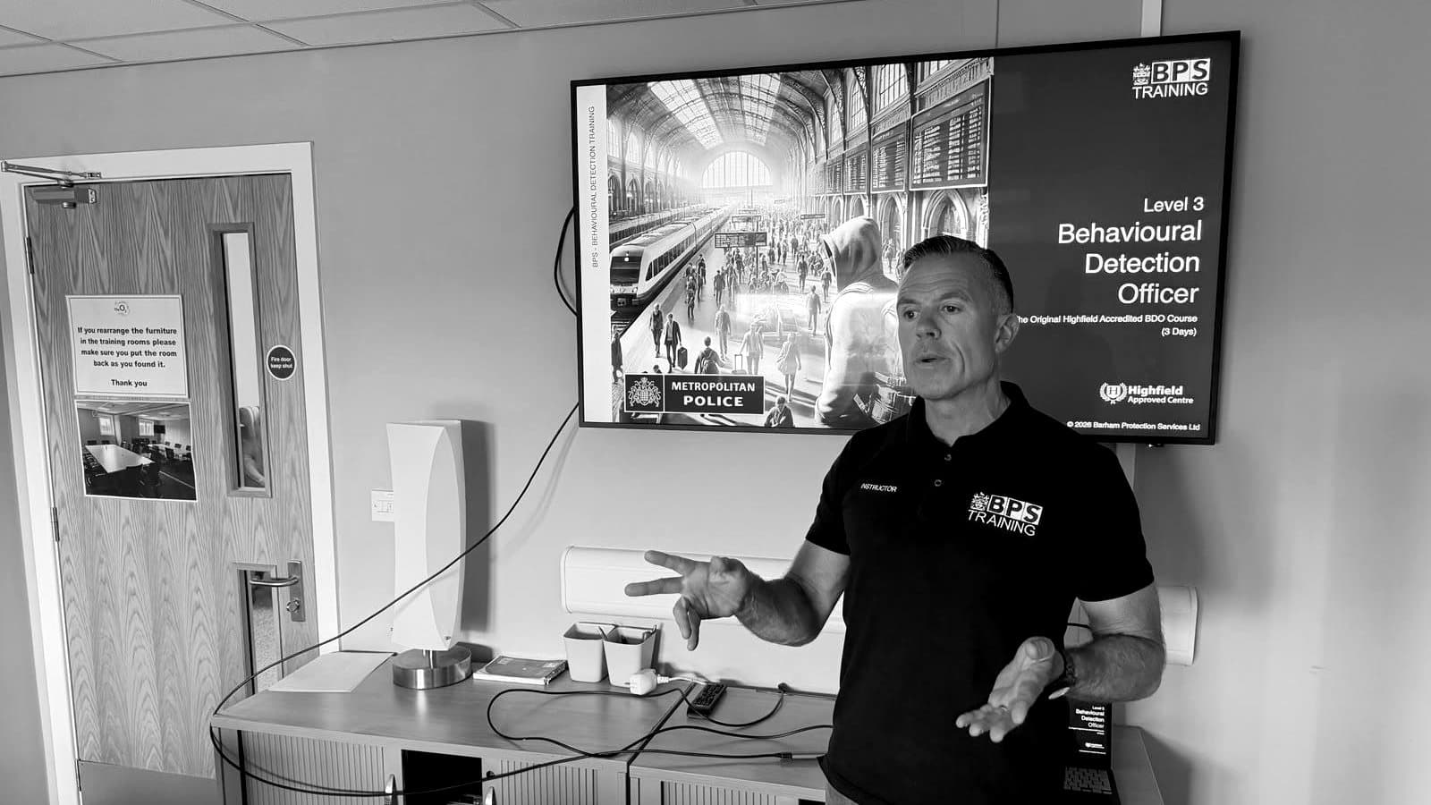 A BPS instructor teaching the Level 3 Behavioural Detection Officer course, the course title on the screen behind him