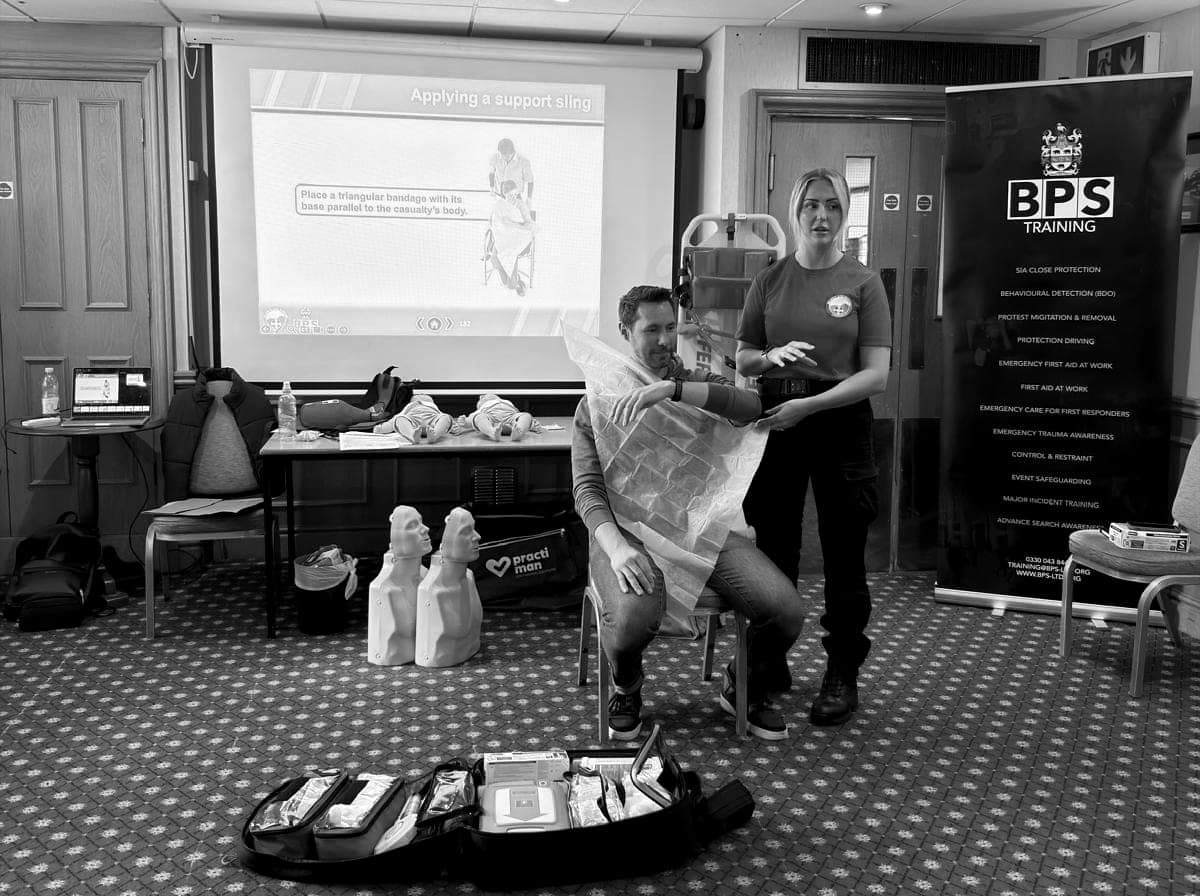 A support sling demonstrated on a seated volunteer at a BPS first aid training session, the slide on screen and the BPS Training banner beside them