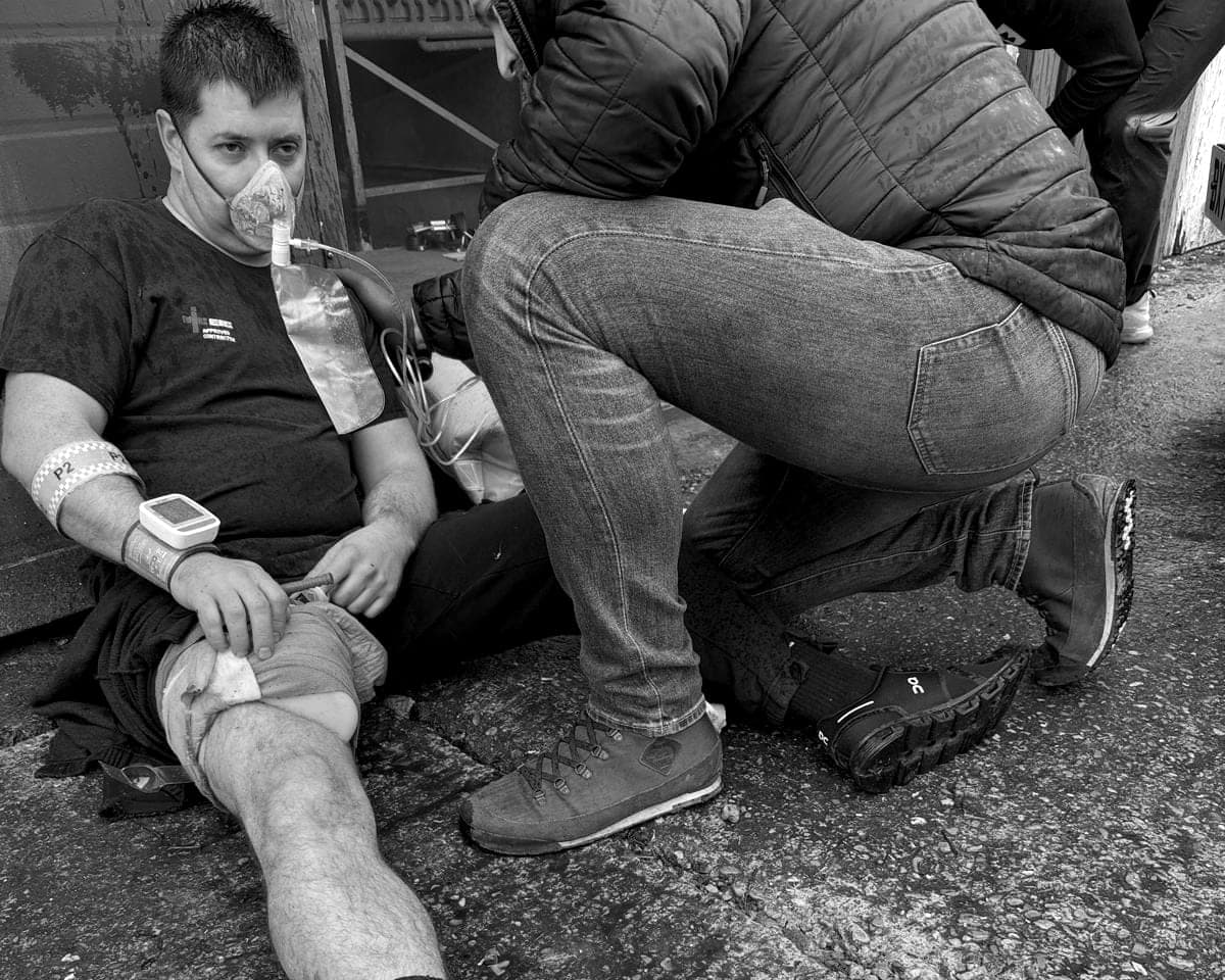 A BPS medical training scenario: a role-play casualty sits on wet ground with an oxygen mask and a make-up leg wound while a course member kneels beside him